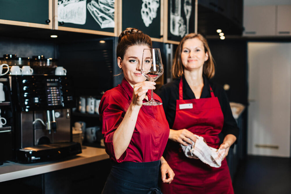 Zwei Damen in Service-Kleidung stehen vor einer Kaffeemaschine und prüfen, ob ein Weinglas sauber geworden ist