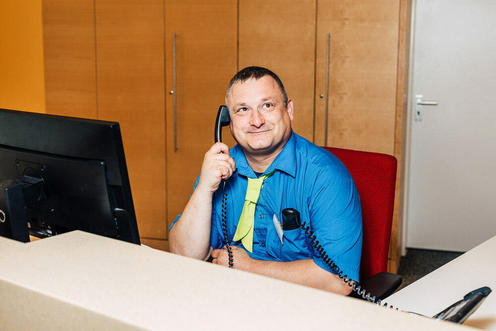 Ein Mann sitzt an einer Hotelrezeption und hat einen Telefonhörer in der Hand 