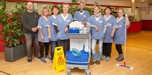 Eine Gruppe von Reinigungsfachkräften der Züssower Service und Catering steht hinter einem Putzwagen mit einem Wischmopp zusammen