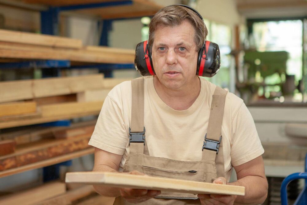 Ein Mann mit Gehörschutz hält ein gefertigtes Brett; hinter ihm ein Holzstapel