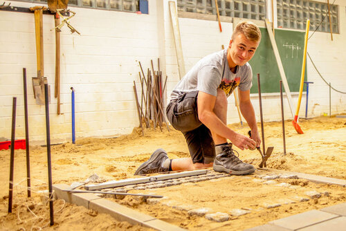 Ausbildung in der Fachrichtung Tiefbau Ein junger Mann hockt auf sandigem Untergrund und hat einen Hammer in der Hand