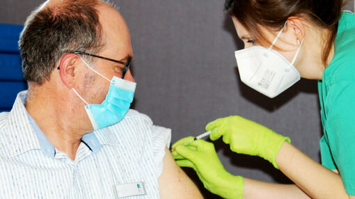 Eine Ärztin in Arztkleidung drückt eine Spritze in die Schulter eines Mannes, der eine Brille und eine Atemschutzmaske trägt