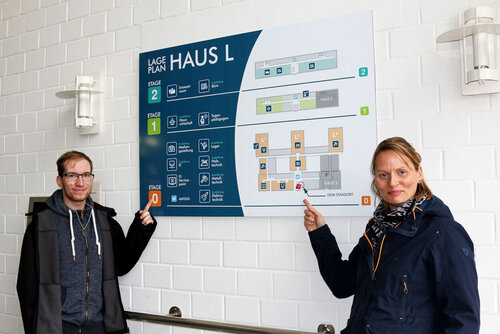 Eine Frau und ein Mann zeigen auf eine große Grafik an einer Wand