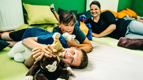 Eine ältere Erzieherin und zwei Kinder liegen mit Kuscheltieren auf dem Boden und spielen