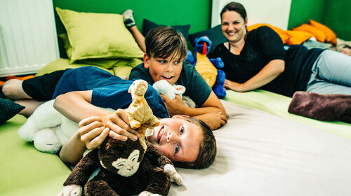 Eine ältere Erzieherin und zwei Kinder liegen mit Kuscheltieren auf dem Boden und spielen