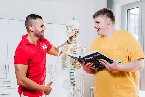 Zwei junge auszubildende Pflegekräfte stehen mit einem Lehrbuch an einem Skelett und lernen Anatomie