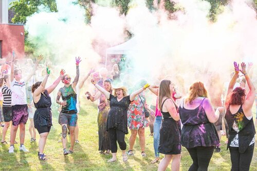 Mehrere Menschen stehen freudig beisammen, strecken die Arme, jubeln und sind in einen Nebel aus Farben gehüllt.
