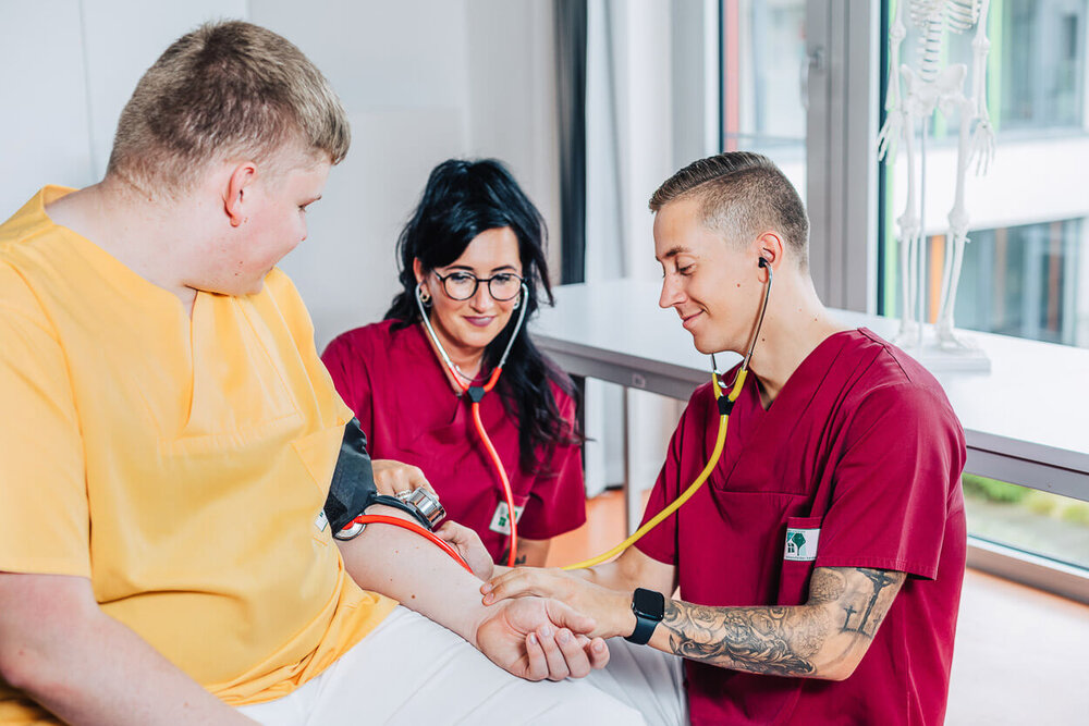 Ein junger Alten- und Krankenpflegehelfer misst einer auszubildenden Kollegin den Blutdruck