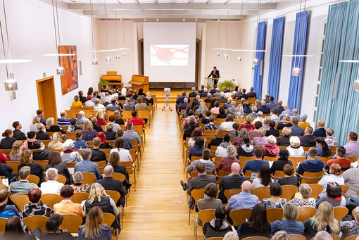 Ein großer Saal mit vielen Menschen, die in Sitzreihen sitzen und am Ende steht ein Mann an einem Pult