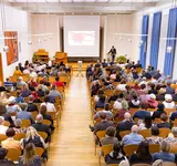 Ein großer Saal mit vielen Menschen, die in Sitzreihen sitzen und am Ende steht ein Mann an einem Pult