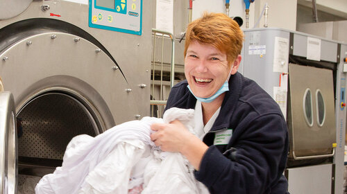 In der Wäscherei der Greifenwerkstatt Eine Frau holt einen großen Berg Wäsche aus einer sehr großen Trockenmaschine