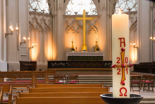 Eine angezündete Kerze vor dem Hintergrund von leeren Kirchenbänken und dem Hochaltars mit Kreuz