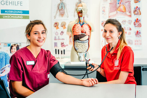 Zwei junge Frauen in Arbeitskleidung sitzen an einem Tisch und die eine misst der anderen den Blutdruck