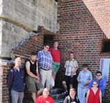 Eine Gruppe Menschen hat sich auf Stufen gesetzt vor einem alten, halb verfallenen großen Industriegebäude aus Backstein